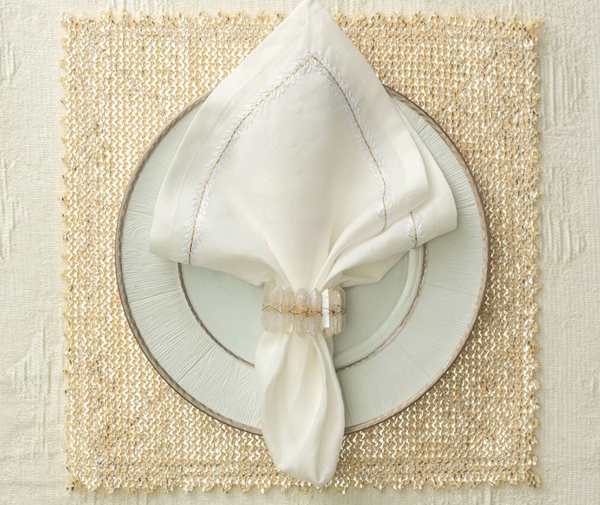 A white cloth napkin with a Kim Seybert Radiant Napkin Ring Iridescent is neatly placed on a white plate with silver edges, resting on a gold woven placemat atop a cream-colored tablecloth.
