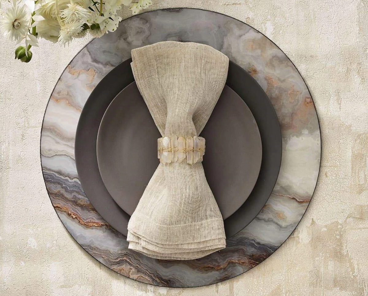 A beige linen napkin with a Kim Seybert Radiant Napkin Ring Iridescent Set of 4 rests on dark gray plates atop a marbled charger, arranged on a textured light surface with white flowers in the corner.