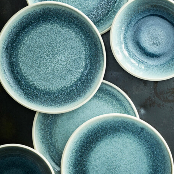Rosenthal Junto Aquamarine Dinnerware plates and bowls feature a crackled blue reactive glaze with varying shades and textures, arranged on a dark surface. Dishwasher and microwave safe for daily use.