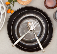 A Marcus Black Dinnerware Set of 4 by Blue Pheasant, with a silver spoon and fork crossed at the center, is paired with organic-shaped dishware, a brown glass, salt, and orange kumquats on a light surface.