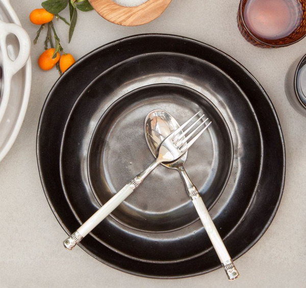 A Marcus Black Dinnerware Set of 4 by Blue Pheasant, with a silver spoon and fork crossed at the center, is paired with organic-shaped dishware, a brown glass, salt, and orange kumquats on a light surface.