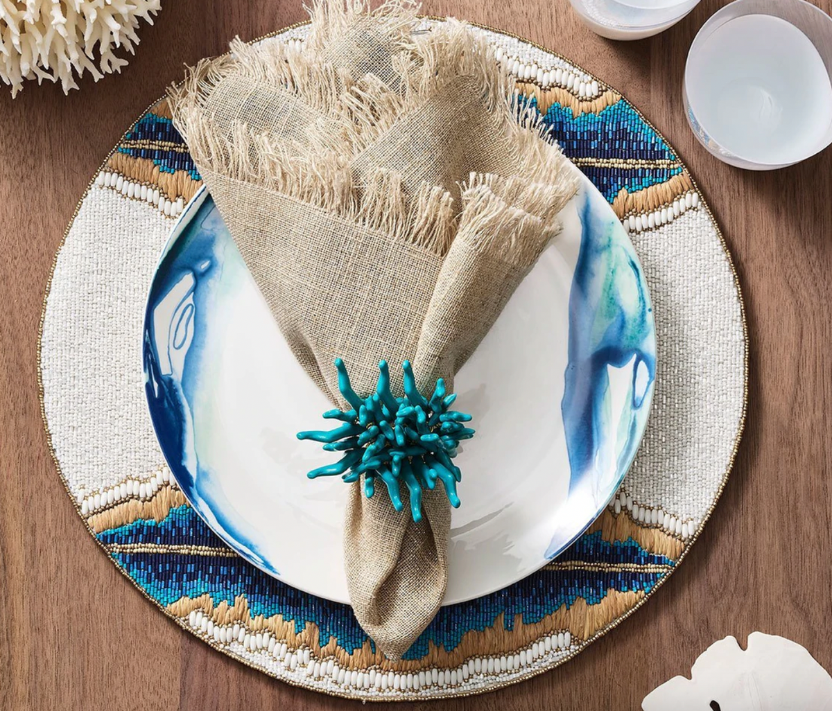 A table setting featuring Kim Seybert’s Fringe Napkin Silver & Natural Set of 4 on a blue and white plate with a beaded placemat, turquoise coral napkin ring, white bowls, and decor atop a wooden table.
