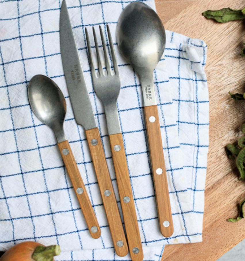 The Sabre Bistro Flatware 5 Piece Setting - Teak, featuring stainless steel utensils with wooden handles, is displayed on a blue and white checkered cloth atop a wooden surface, with onion and leafy greens at the edges.