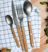 The Sabre Bistro Flatware 5 Piece Setting - Teak, featuring stainless steel utensils with wooden handles, is displayed on a blue and white checkered cloth atop a wooden surface, with onion and leafy greens at the edges.