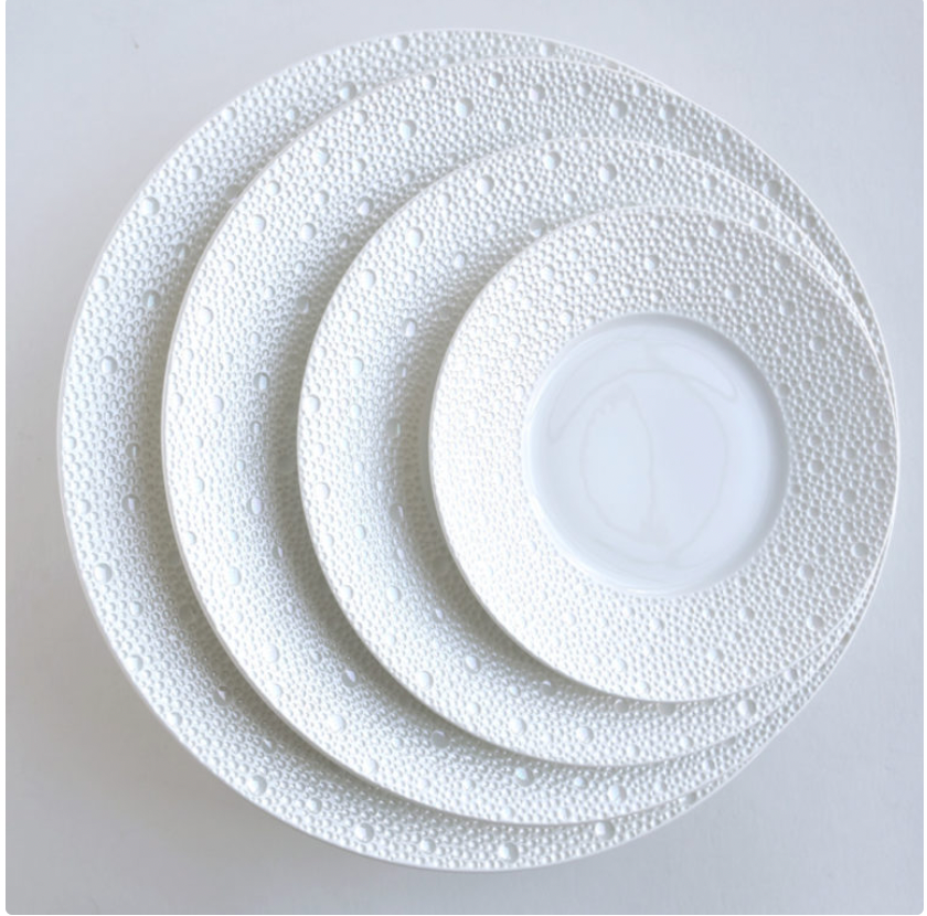 Four white Ecume Dinnerware Perle plates by Bernardaud are stacked on a flat, light surface. The rims have a raised pebble-like texture and subtle mother-of-pearl finish, while the centers are smooth.
