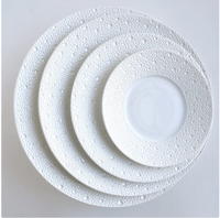 Four white Ecume Dinnerware Perle plates by Bernardaud are stacked on a flat, light surface. The rims have a raised pebble-like texture and subtle mother-of-pearl finish, while the centers are smooth.