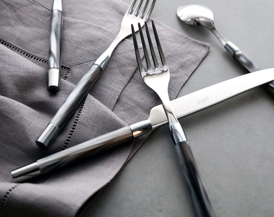 A close-up of the Capdeco Conty Large Serving Spoon Grey, French-made from stainless steel, with a dark glossy handle, arranged on a gray cloth napkin atop a gray surface.