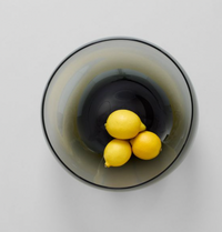 A top-down view of three yellow lemons in the Gary Bodker Nesting Glass Bowl Charcoal, arranged on a light gray surface to showcase the bowl’s handcrafted elegance.