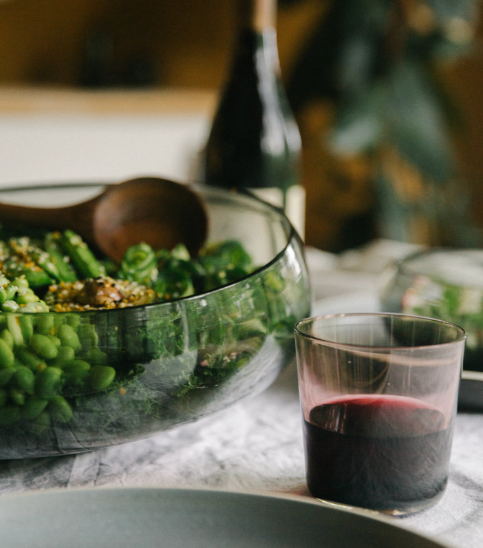 The Gary Bodker Nesting Glass Bowl Charcoal, filled with green salad and paired with wooden utensils, sits beside a glass of red wine on the table; a bottle is softly blurred in the background.