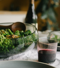 The Gary Bodker Nesting Glass Bowl Charcoal, filled with green salad and paired with wooden utensils, sits beside a glass of red wine on the table; a bottle is softly blurred in the background.