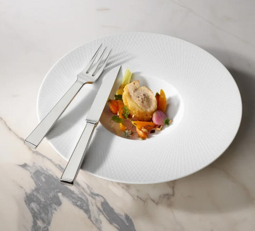 A small gourmet dish with sliced meat and vegetables is served on a large white plate, accompanied by the Degrenne Squado Flatware 5 Piece Setting on a marble surface.