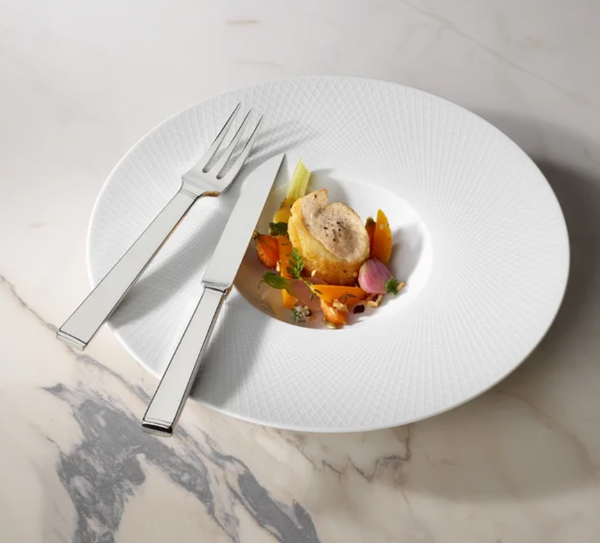 A small gourmet dish with sliced meat and vegetables is served on a large white plate, accompanied by the Degrenne Squado Flatware 5 Piece Setting on a marble surface.