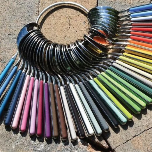 The Mepra Fantasia Flatware 5 Piece Set, shown in a semicircle with vibrant resin handles in various colors, is displayed on a textured stone surface.