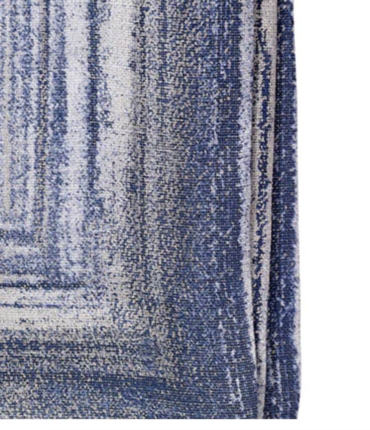 A close-up of the Bodrum Cornice Napkin Navy Set of 4 shows a textured blue and white napkin with a faded gradient pattern, visible woven fibers, and a subtle metallic border along one edge, slightly folded over.