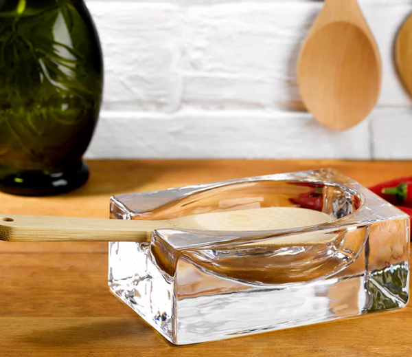 The Nude Glassware Glass Modern Spoon Rest holds a wooden spoon on a countertop, with kitchen utensils and a green vase in the background against a white brick wall.