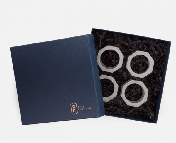 A navy Blue Pheasant box sits open, revealing four Arthur Leather Grey napkin rings nestled in black crinkle paper—a touch of luxury table decor for any setting.