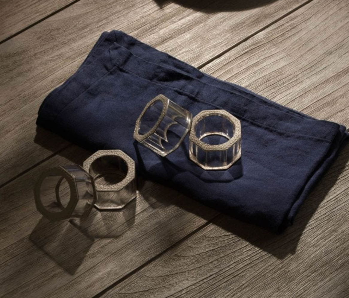 The Arthur Leather Grey Napkin Ring Set of 4 by Blue Pheasant rests on a folded dark blue napkin atop wood, with soft lighting and gentle shadows—an elegant touch for your luxury table decor.
