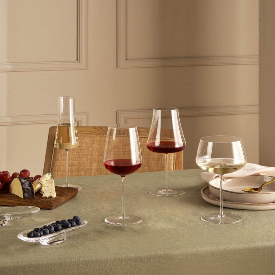 A table set with three Nude Glassware Stem Zero Barware wine glasses, a champagne flute, cheese and grapes, blueberries in a dish, and stacked plates with a gold spoon on a beige tablecloth.