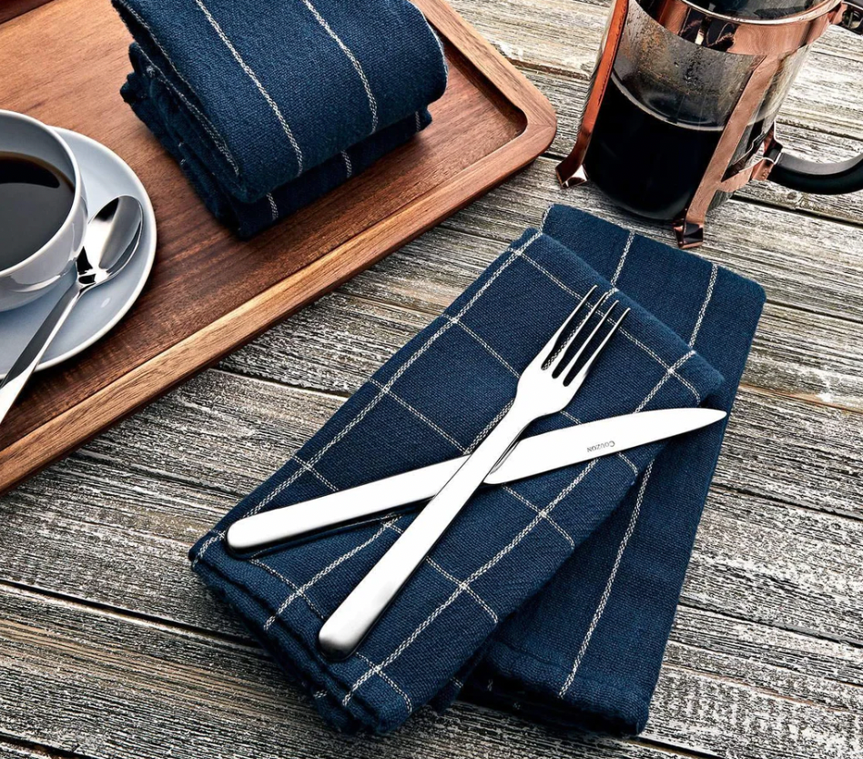 A folded blue checkered napkin with Couzon/DeVine Corp /MAISON3AMIS Alkaline Flatware 5 Piece Setting - Stainless Steel rests on a wooden table, beside a cup of coffee, French press, extra napkin, and a wooden serving tray.
