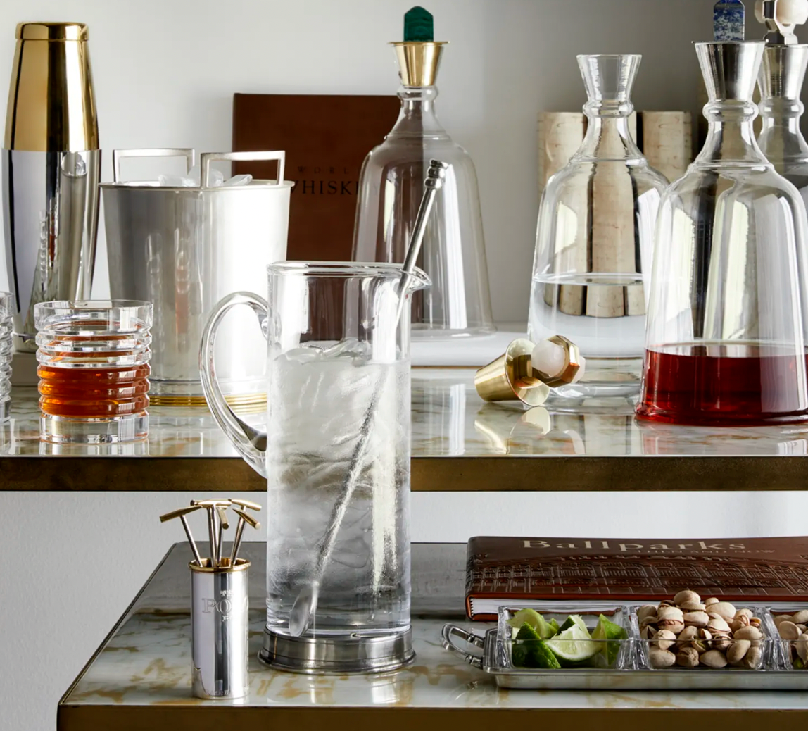 A marble bar cart displays liquor decanters, the MATCH Martini Crystal Pitcher by MATCH, glasses, an ice bucket, a handmade pewter coaster set, lime wedges and pistachios on a tray, and cocktail picks on a steel surface.