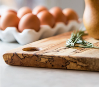 Close-up of the Peterman Spalted Maple Live Edge Board 21" with a sprig of rosemary; brown eggs sit in a white ceramic holder in the background.