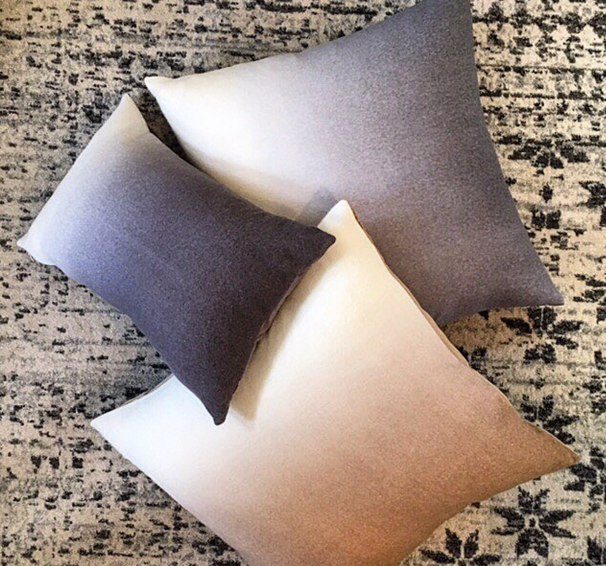 The Johanna Howard Dip Dye Pillow in Light Grey is arranged with two other gradient-toned pillows on a neutral, textured patterned rug.