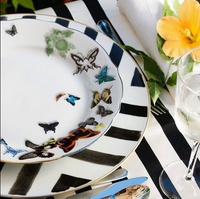 A luxurious table setting with a white Vista Alegre Butterfly Parade Dinnerware plate adorned with colorful butterflies, placed on a black-and-white striped charger, alongside cutlery, a yellow flower, and sparkling water.