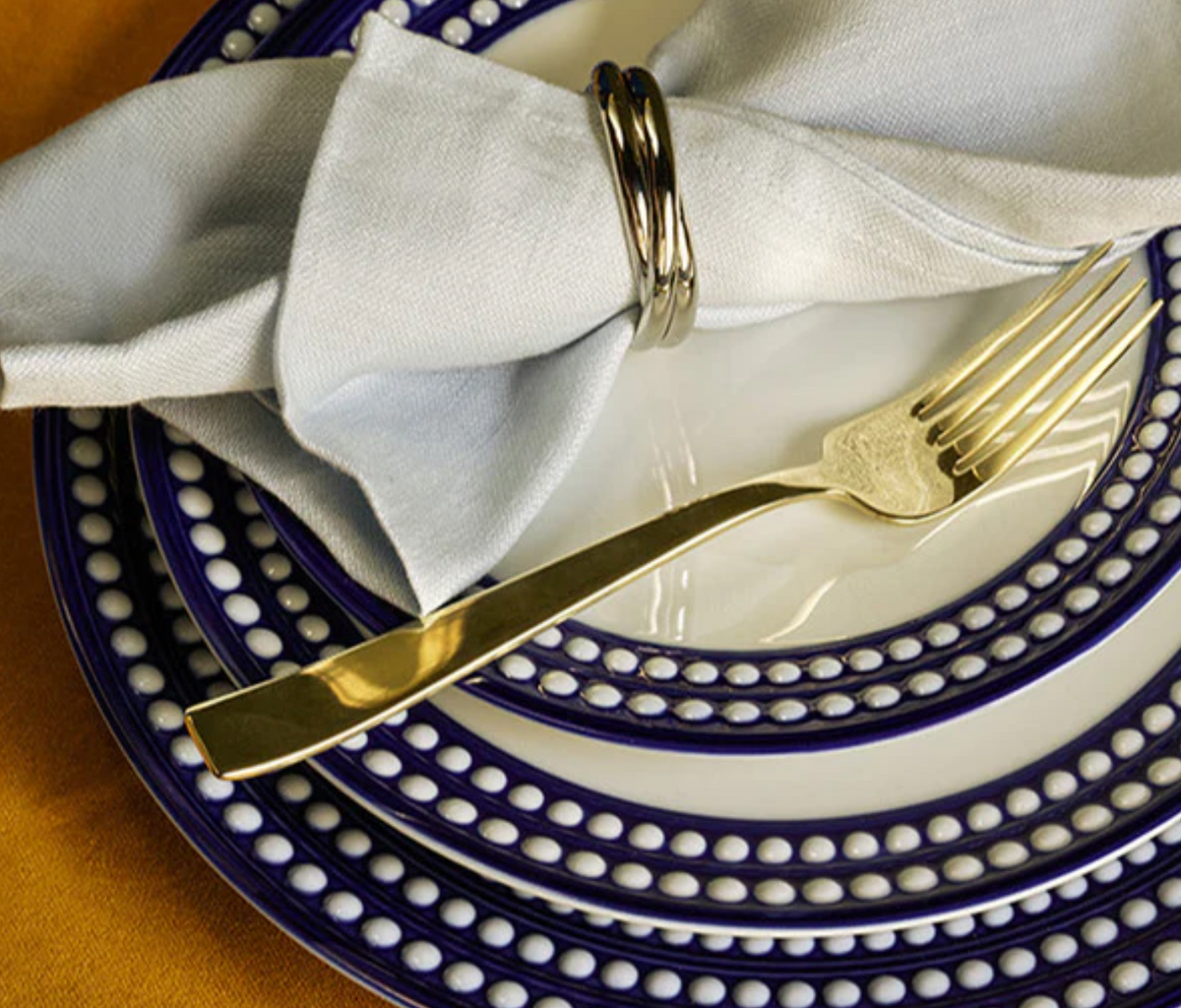 A gold fork rests on a L'Objet Perlèe Blue Dinnerware plate with blue dotted trim, topped by a folded light gray napkin in a silver ring, set on a brown or gold tablecloth.