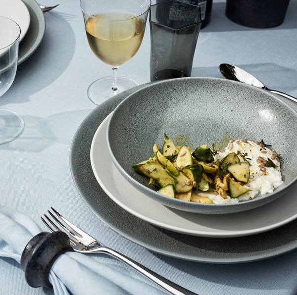 A bowl of zucchini and ricotta sits on stacked L'Objet Terra Seafoam Dinnerware beside a fork, a napkin with a ring, white wine glass, and water glass atop a light blue tablecloth.