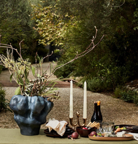 A table set outdoors features the Timna Vase Short by L'Objet holding branches, a gold candelabra with white candles, bottles, glasses, fruit, crackers, and napkins amid greenery and a tree-lined path in the background.