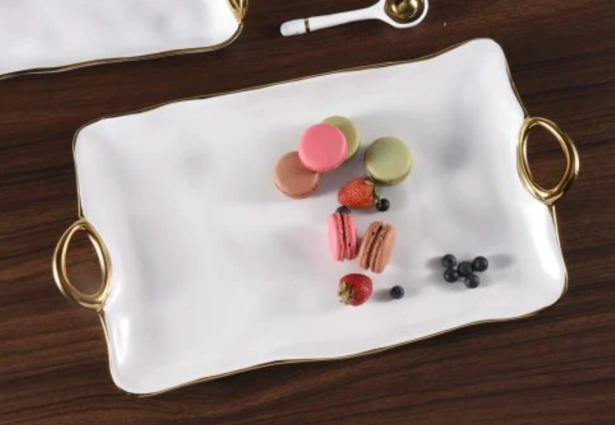 The PAMPA BAY Gold Handle Tray Large, made of white porcelain with gold handles and dishwasher safe, holds macarons, strawberries, and blueberries. A small spoon and another dish are partially visible on the dark wooden surface nearby.