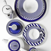 The Harvard Dinnerware set by Vista Alegre features bold blue and white geometric patterns on plates, cups, a teapot, and a tall pot. Made in Portugal, the pieces are arranged neatly on a white background.