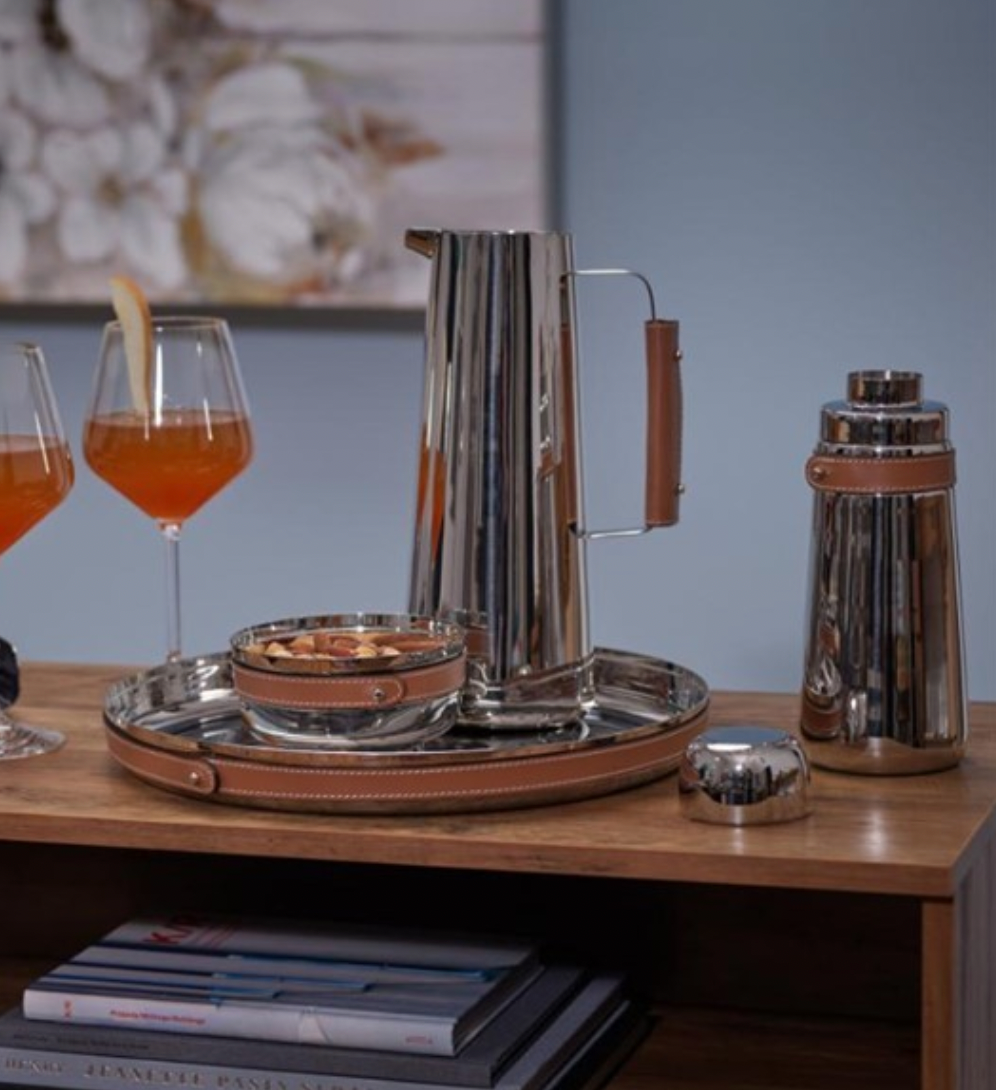 A polished metal tray with leather handles holds modern barware—a matching pitcher, stainless steel tools, two orange cocktails, and the Nambe Tahoe Nut Bowl—arranged on a wooden table with books and a metal shaker nearby.