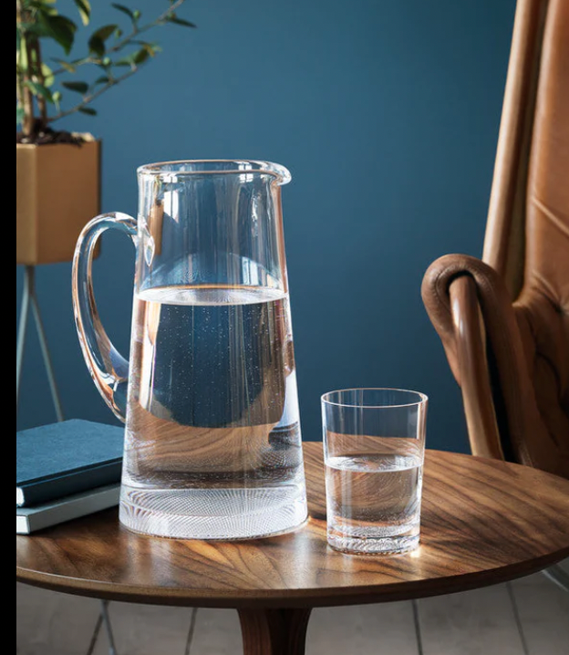 An Orrefors Kosta Boda Limelight Jug and a crystal glass of water rest on a round wooden table by a closed blue notebook, with a leather chair and potted plant against a blue wall in the background.