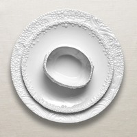 Three Haas Mojave White Dinnerware pieces by L'Objet, featuring textured bubble and swirl patterns, are stacked on a neutral fabric; the top dish displays an irregular, organic shape.