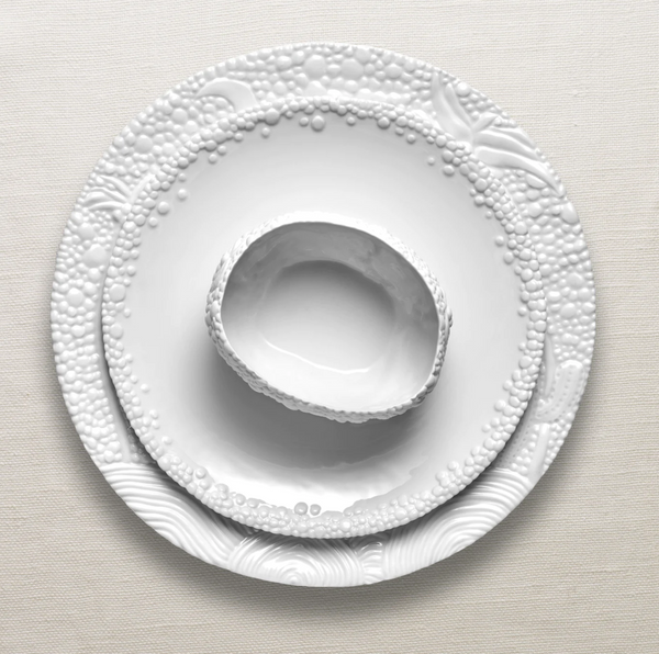 Three Haas Mojave White Dinnerware pieces by L'Objet, featuring textured bubble and swirl patterns, are stacked on a neutral fabric; the top dish displays an irregular, organic shape.
