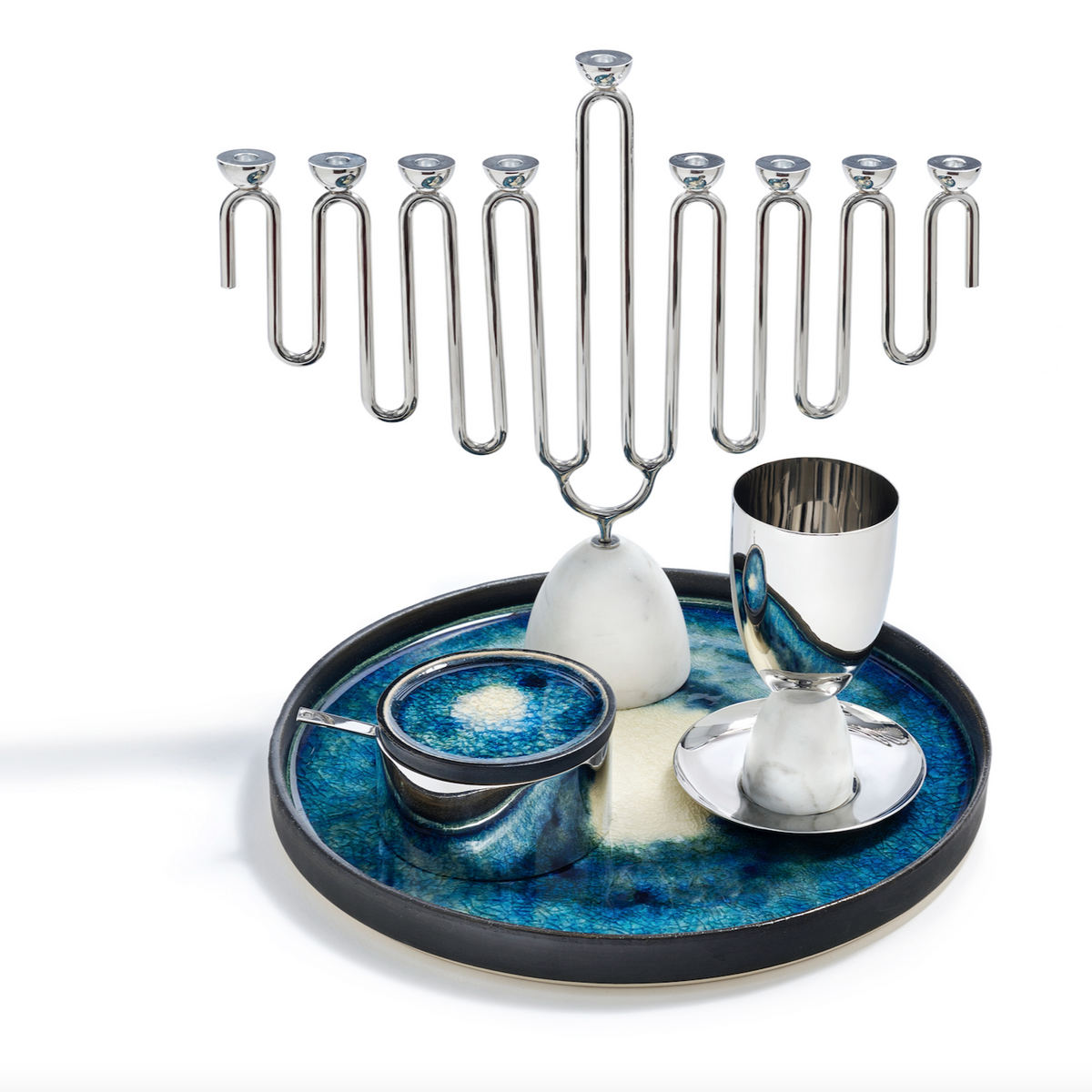 A Coluna Menorah Carrara Marble & Silver by Anna New York stands alongside a goblet and spice container on a round blue and white tray featuring an abstract design. The sleek marble and silver accents add contemporary elegance to the display, making it ideal for any modern celebration.