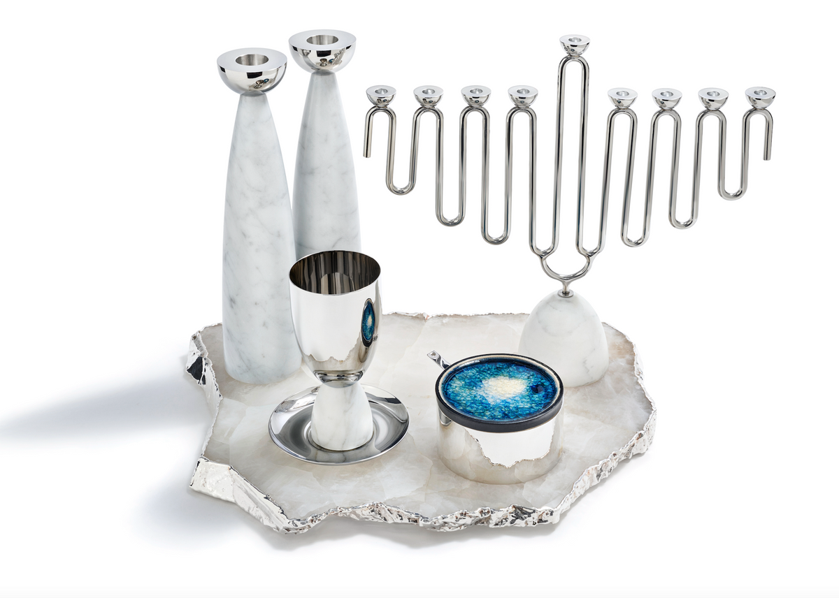 The Coluna Menorah Carrara Marble & Silver by Anna New York, accompanied by two elegant marble candlesticks, a silver goblet, and a marble bowl filled with blue salt, are arranged on a jagged silver tray against a white background.