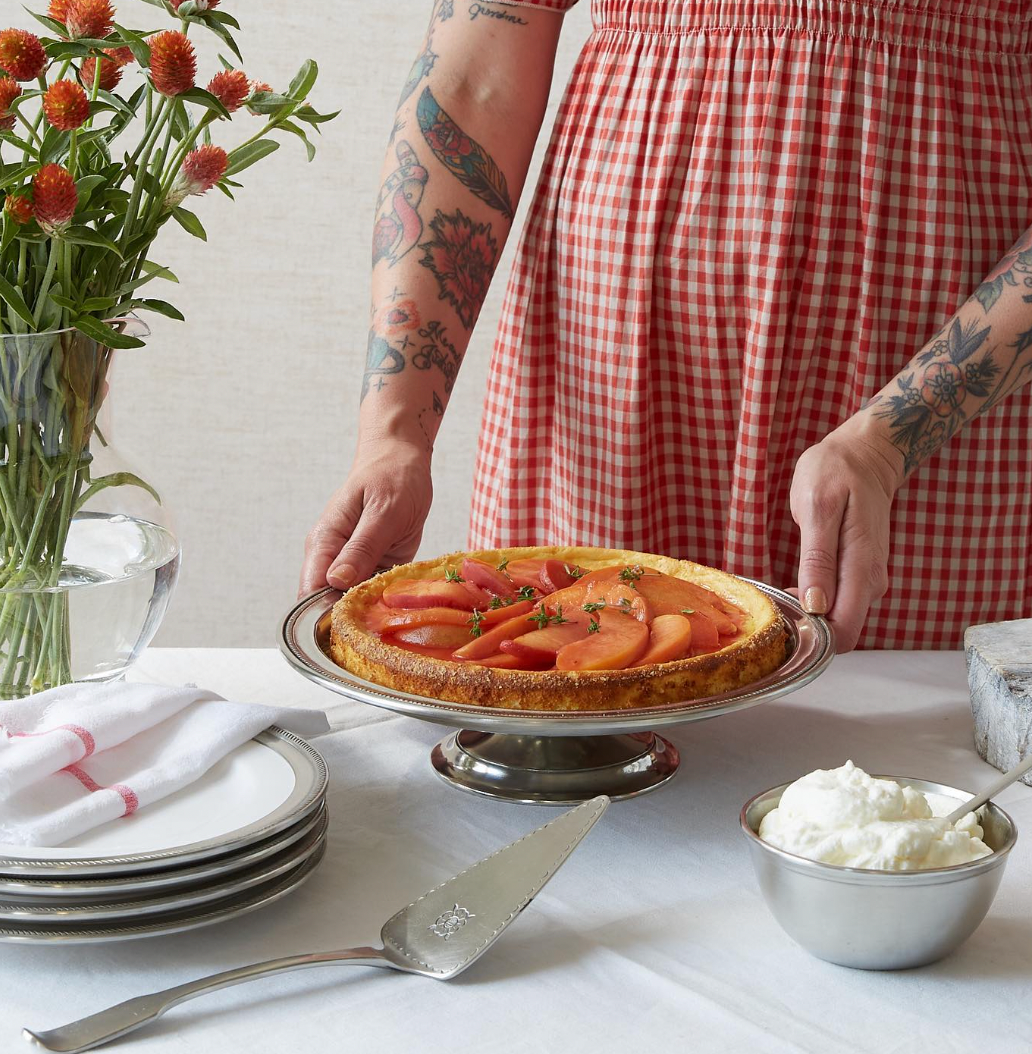 Wearing a red gingham dress with arm tattoos, a person sets a peach-topped tart on MATCH's Convivio cake stand. MATCH Convivio pewter-rimmed plates, pie server, and accessories sit nearby on a white tablecloth with orange flowers.