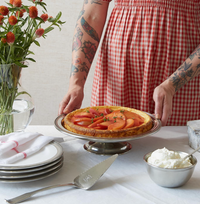Wearing a red gingham dress with arm tattoos, a person sets a peach-topped tart on MATCH's Convivio cake stand. MATCH Convivio pewter-rimmed plates, pie server, and accessories sit nearby on a white tablecloth with orange flowers.
