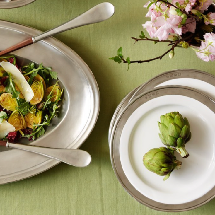 A table set with artichoke salad on a silver platter, two artichokes on MATCH Convivio Italian ceramic dinnerware by MATCH, and a bouquet of pink flowers.