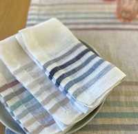 Four neatly folded Bodrum Logan Napkin Blue napkins, featuring pastel and navy stripes, rest on a gray plate atop a matching tablecloth on a wooden table, creating an elegant and calming table setting.