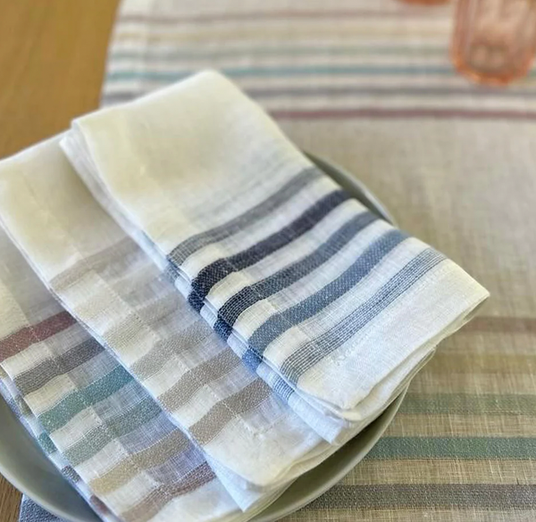 Four neatly folded Bodrum Logan Napkin Blue napkins, featuring pastel and navy stripes, rest on a gray plate atop a matching tablecloth on a wooden table, creating an elegant and calming table setting.