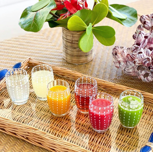 Two Zafferano Perle Beverage Glasses are arranged on a wicker tray atop a woven table, with a green leafy plant in a vase and a purple decorative object in the background.