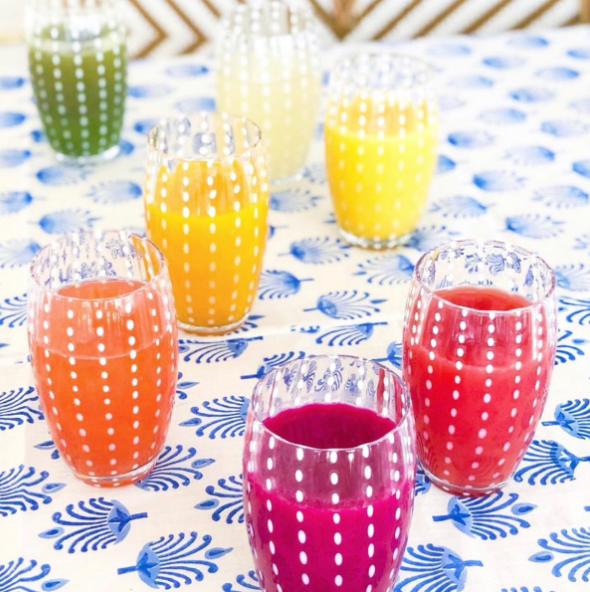 Seven Perle Beverage Glasses by Zafferano, each filled with colorful juice, sit on a white and blue patterned tablecloth; their handmade design features white dotted lines inspired by Federico de Majo’s glassware.