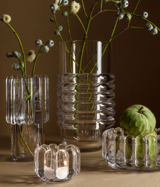 Four Tom Dixon Press Stem Vases by Tom Dixon are grouped together; some elegantly hold white flowers and green stems, one contains green fruit, and a small vase displays two tealight candles. The background is neutral brown.