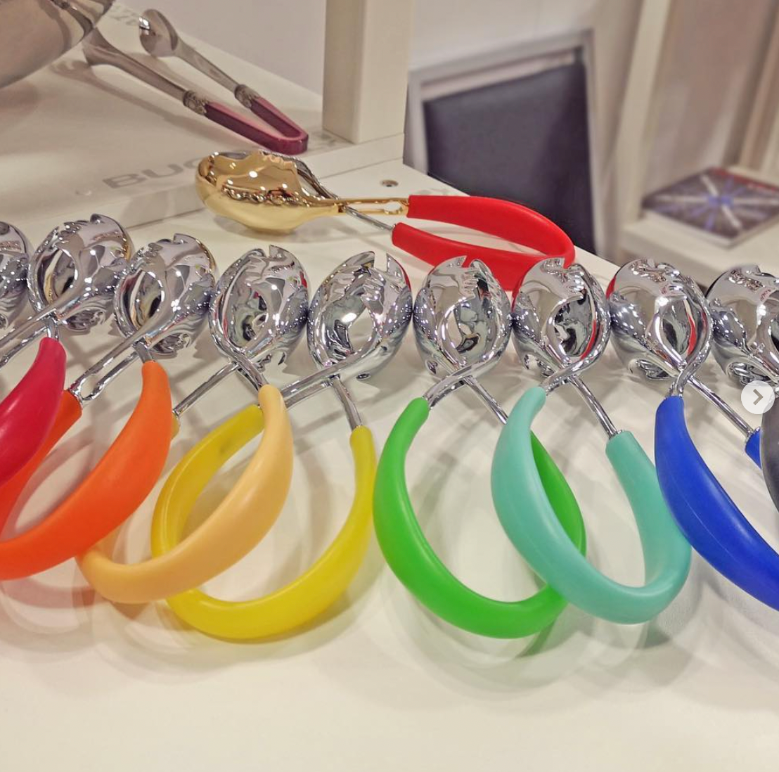A row of Casa Bugatti KISS Salad Tongs in shiny metallic finishes with colorful handles—orange, yellow, green, blue, and red—are displayed on a white surface; a gold tong with red handles is in the background.