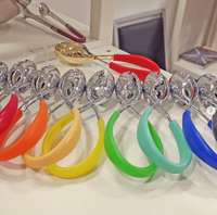 A row of Casa Bugatti KISS Salad Tongs in shiny metallic finishes with colorful handles—orange, yellow, green, blue, and red—are displayed on a white surface; a gold tong with red handles is in the background.