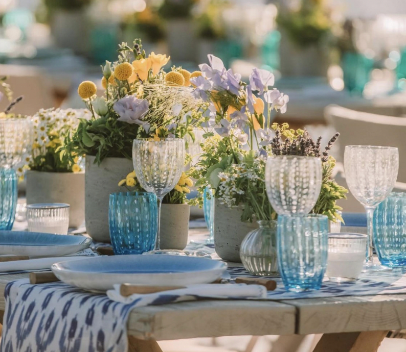 A sunlit outdoor table features Zafferano Perle Wine Goblet Set of 2, white plates, and floral centerpieces with yellow, purple, and white blooms. A blue-patterned runner adds to the relaxed elegance. Made in Italy.