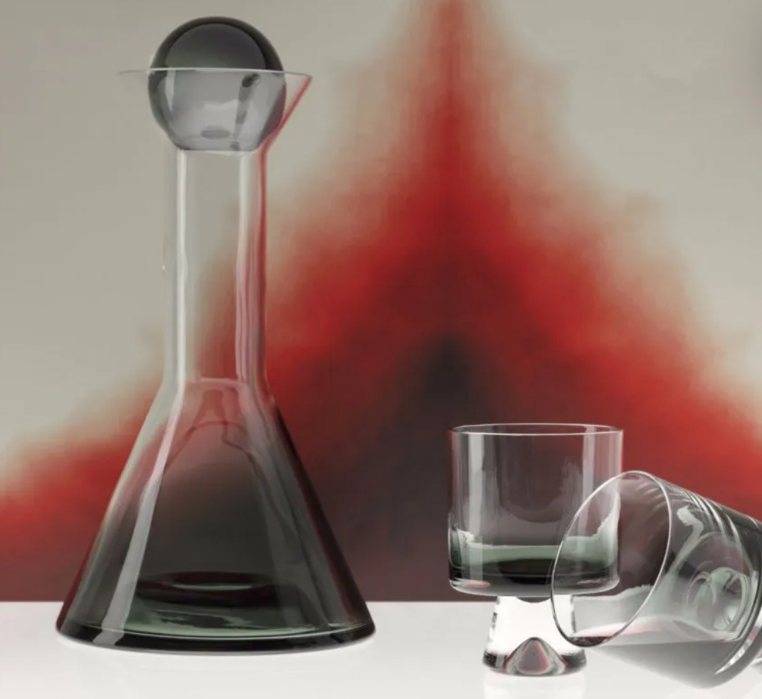 The Tom Dixon Tank Black Decanter, a mouth-blown handmade glass decanter with a spherical stopper, is displayed beside two glasses on a white surface against a bold red abstract background.